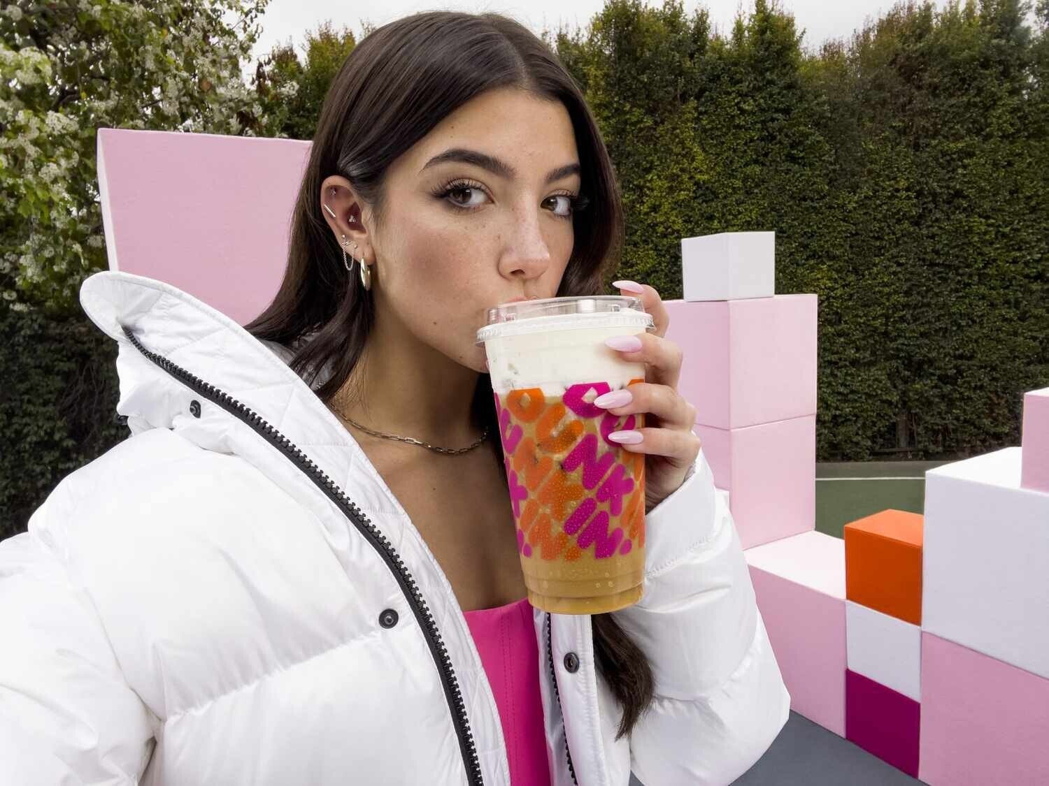 A young woman with long brown hair is pictured wearing a white winter coat and sipping from a large Dunkin&rsquo; Donuts iced coffee.&nbsp;
